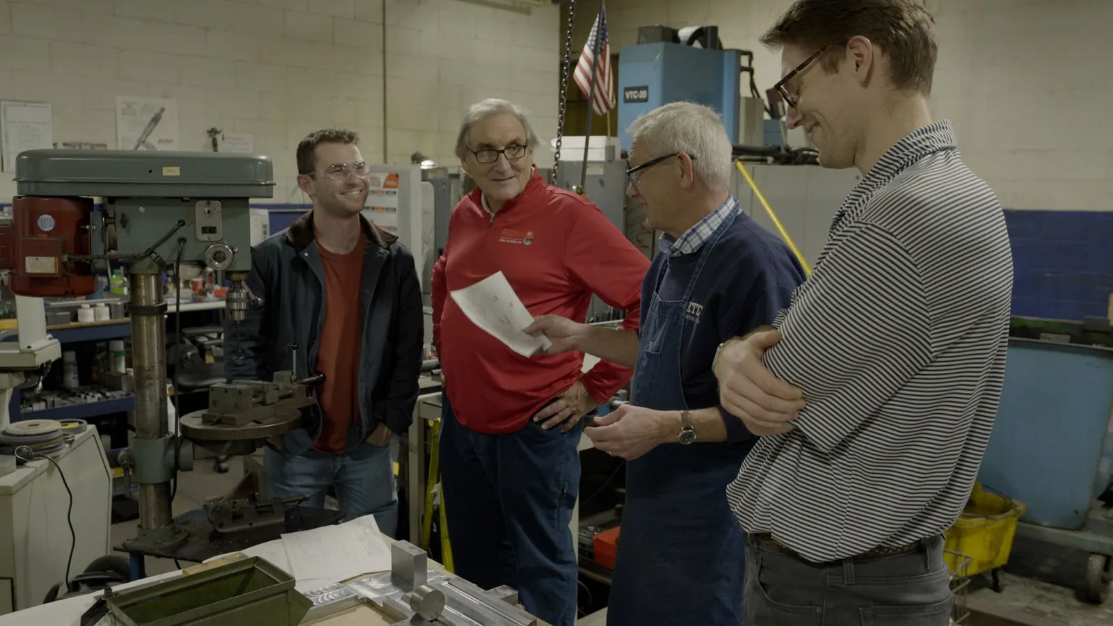 Four men talking in a machine shop, one in a red shirt holding papers