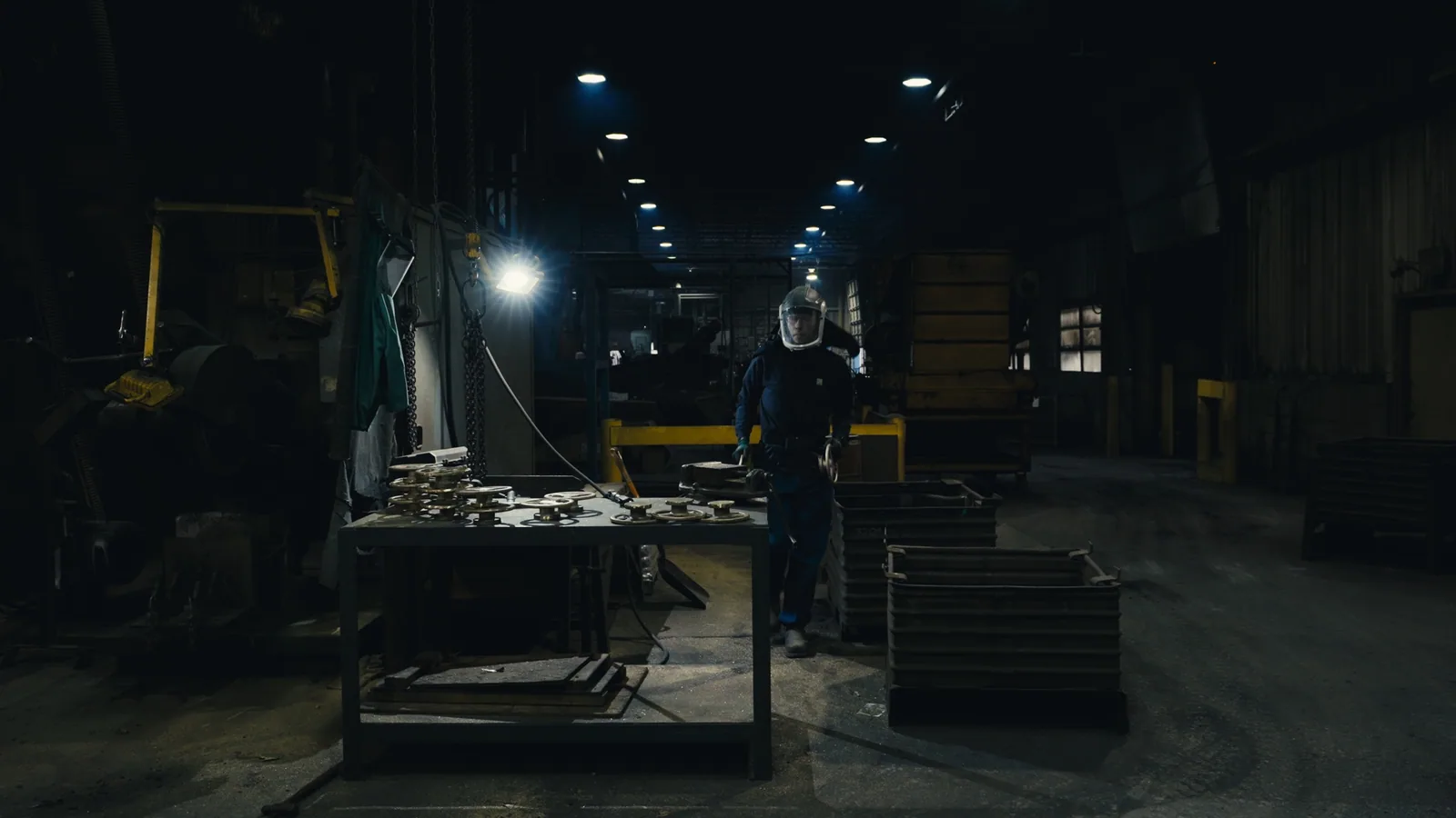 A worker in protective gear crossing a dim foundry floor past racks of castings