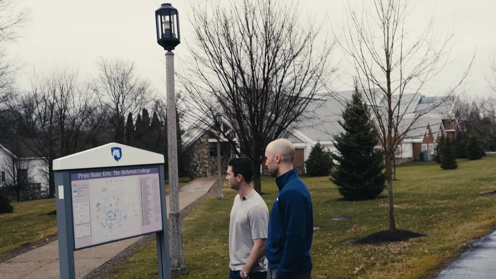 Two men talking on the Penn State Behrend campus beside a map sign, bare winter trees behind