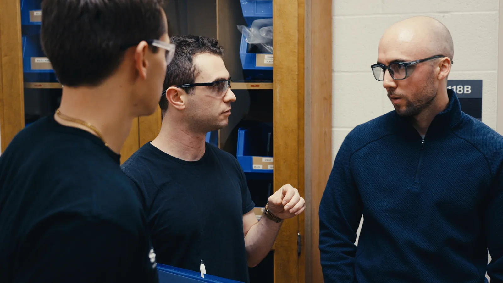 Three men in safety glasses talking in a workshop lined with parts bins