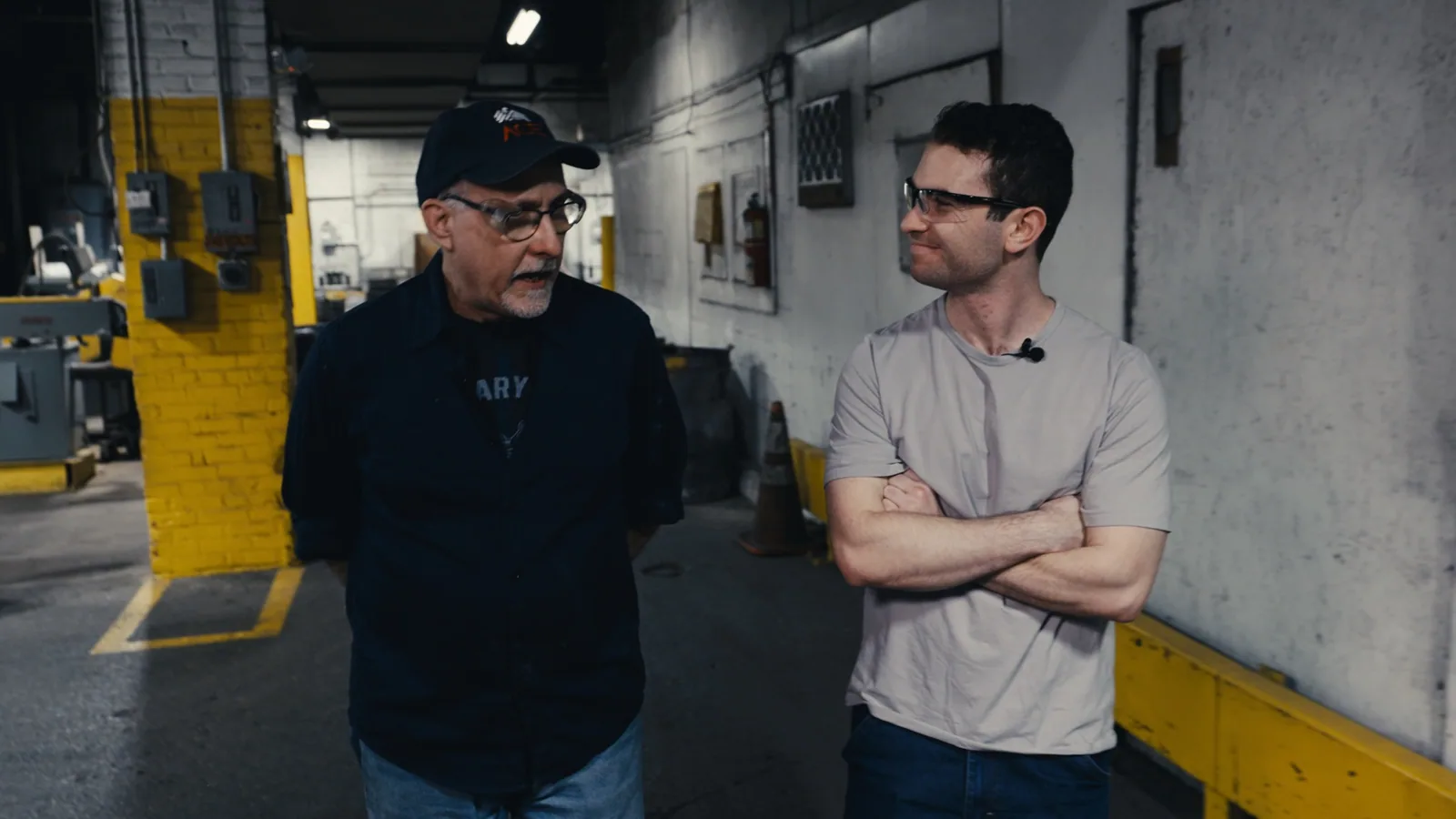 Two men standing and talking on a shop floor, one with arms crossed
