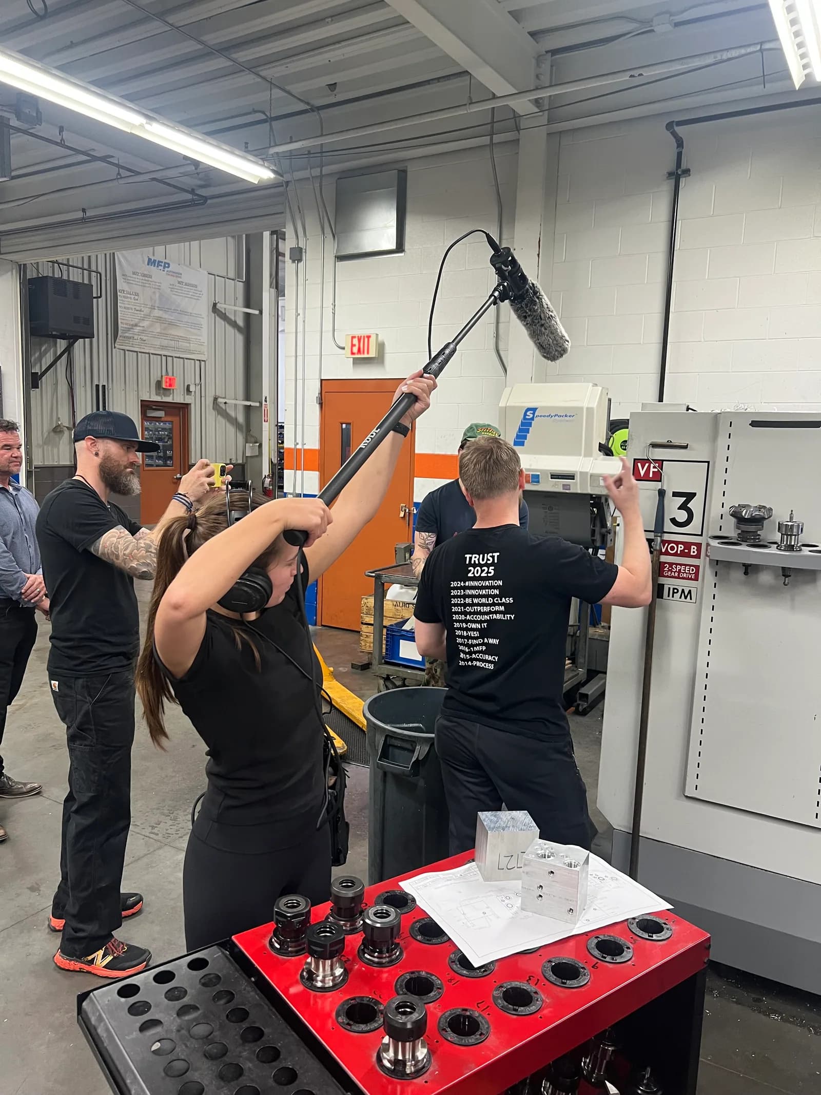 Three people filming beside a CNC machine, one holding a boom microphone overhead