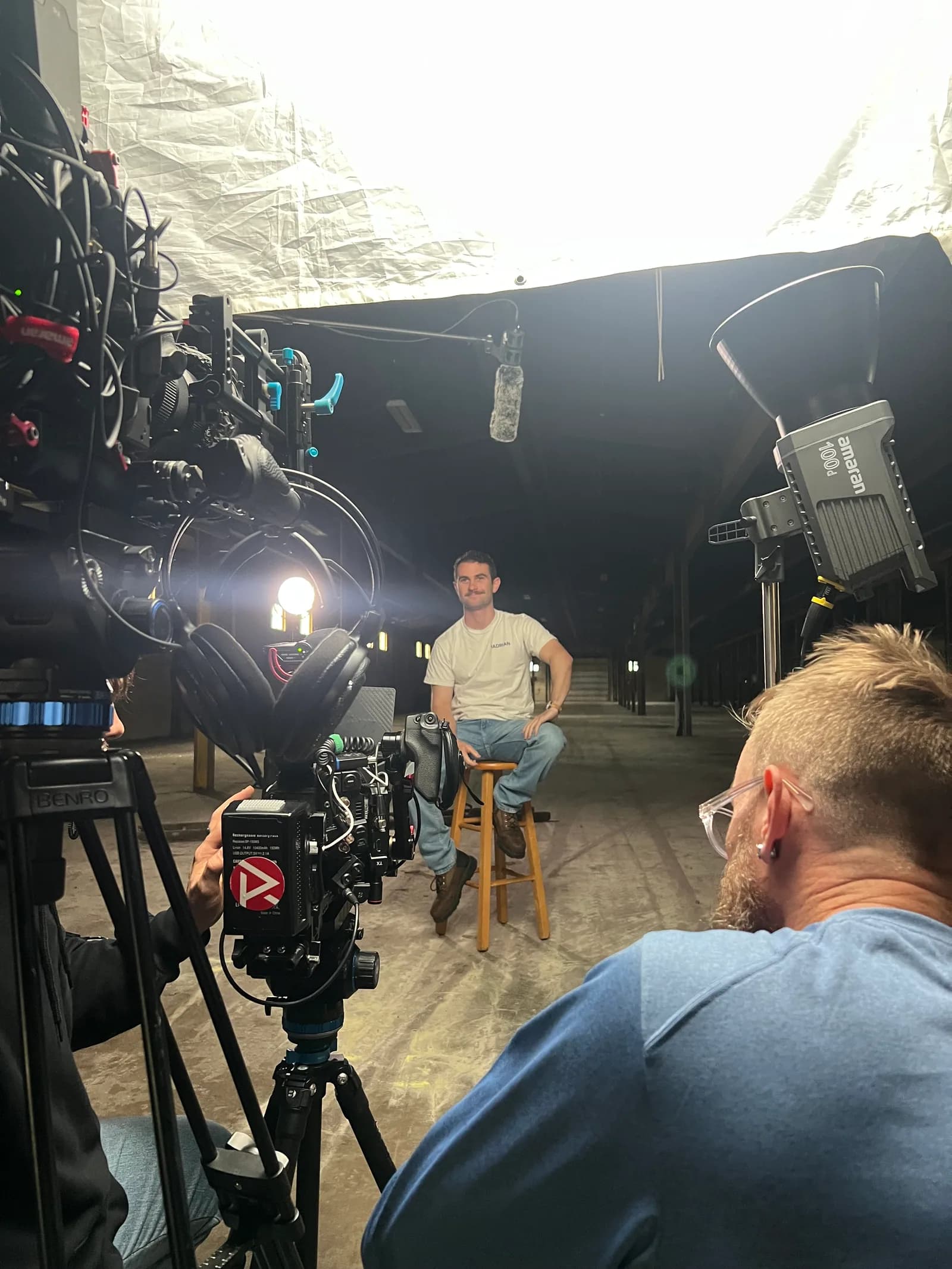 A camera rig and lights trained on a man seated on a stool for an interview inside a factory