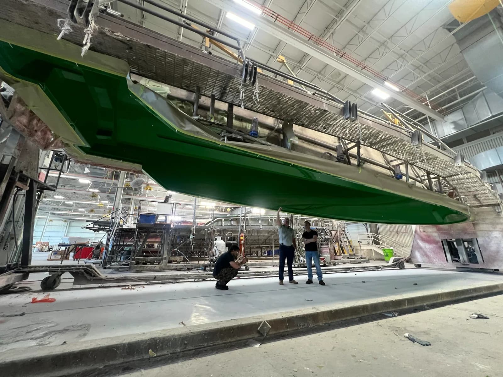 The green hull of a yacht suspended overhead in a boatbuilding hall, workers below