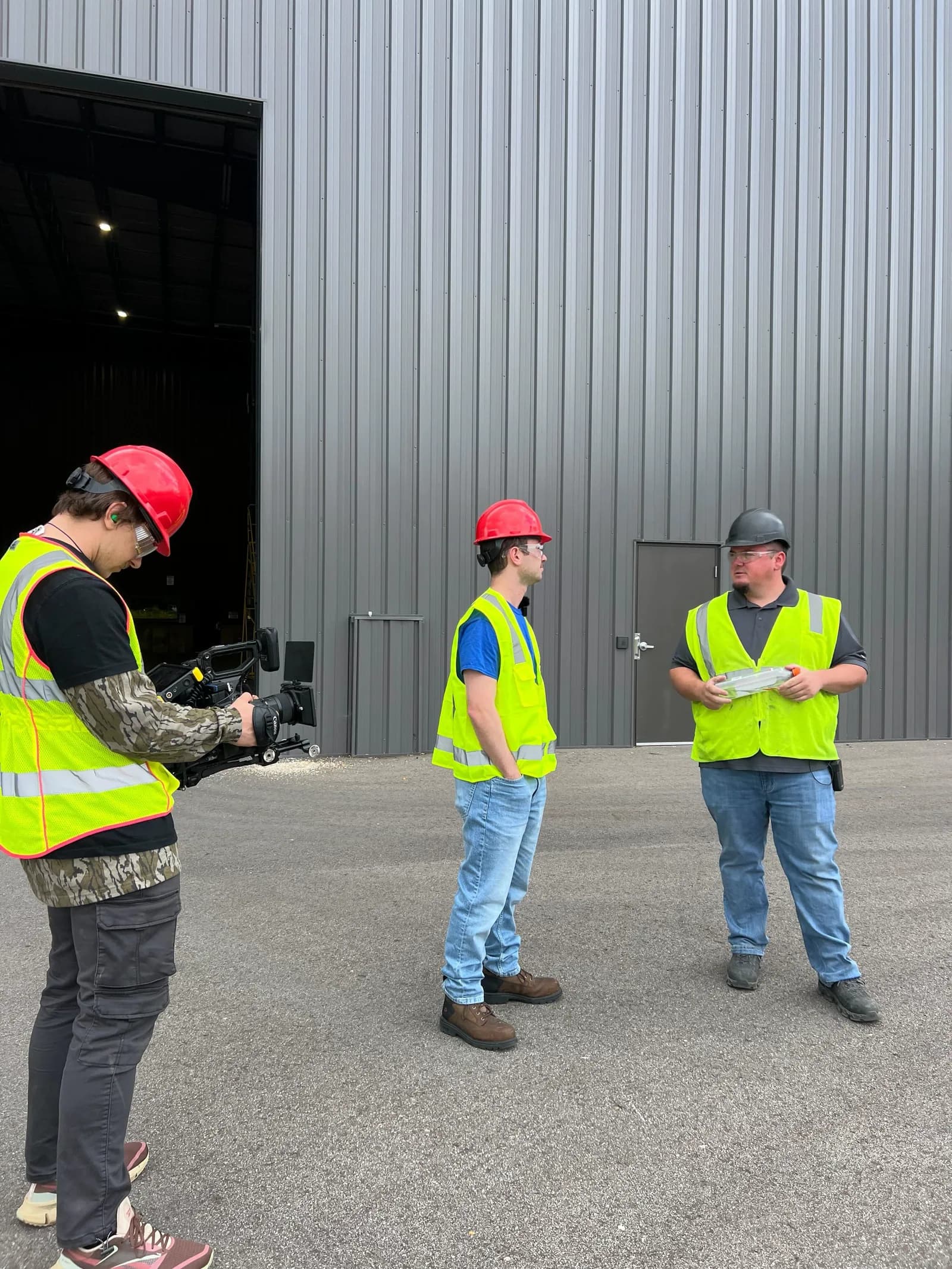 Three men in hard hats and hi-vis vests talking outside a metal building