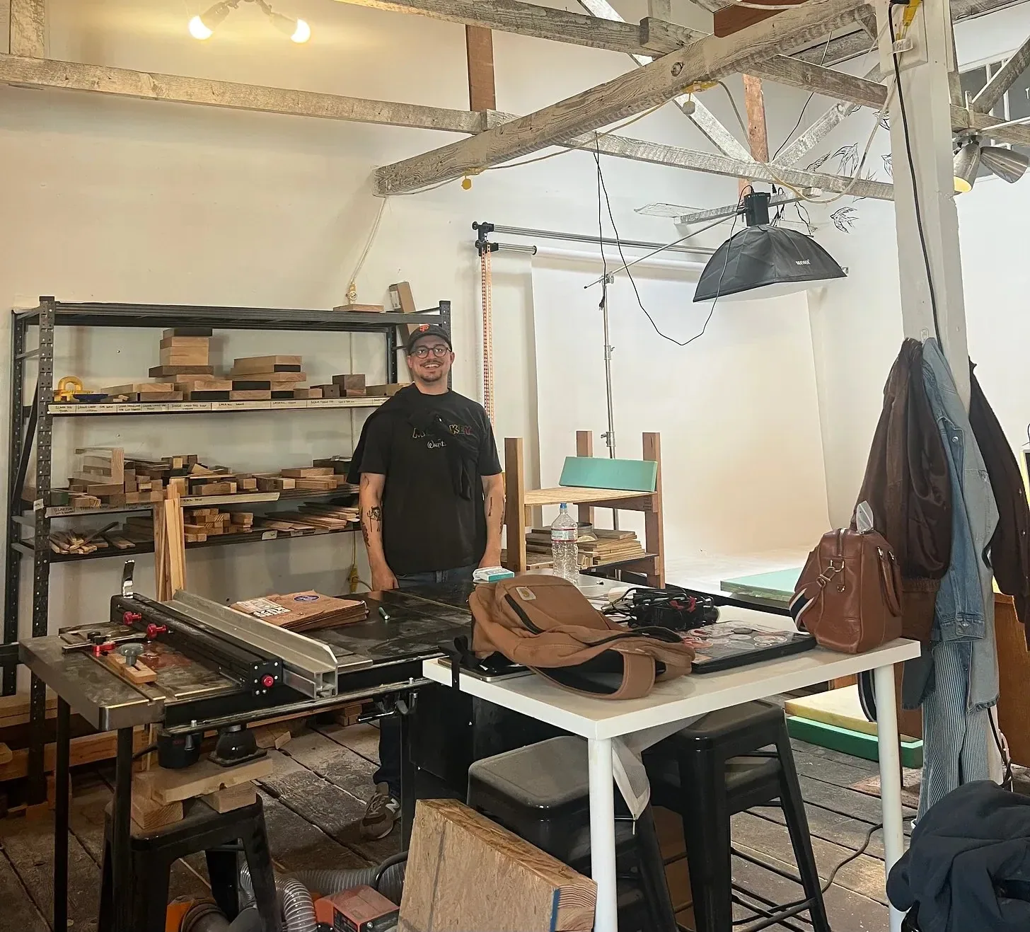 A maker standing in a woodworking shop beside a table saw, lumber racked along the wall and a photography backdrop behind him